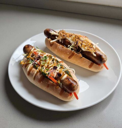 Een bord met daarop 2 hotdogs gemaakt met braadworst.